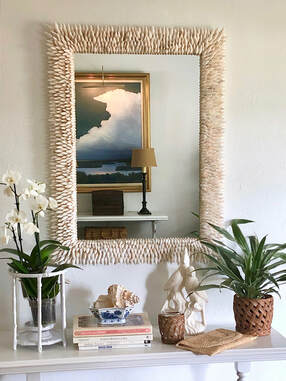 A white mirror made with pointy white seashells is hung above a table with orchids and books.