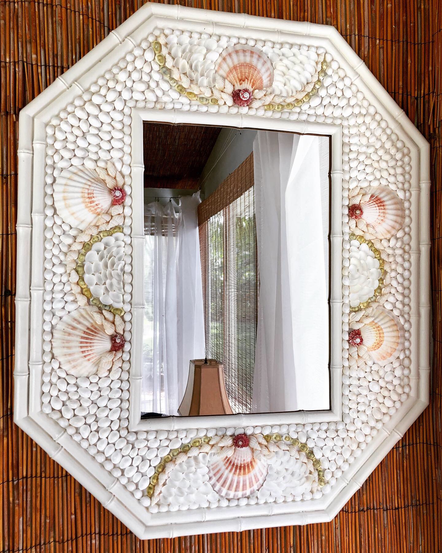 Mirror made from faux bamboo is painted white and has lots of shells placed in bridged patterns. It reflects a window with shear white curtains.
