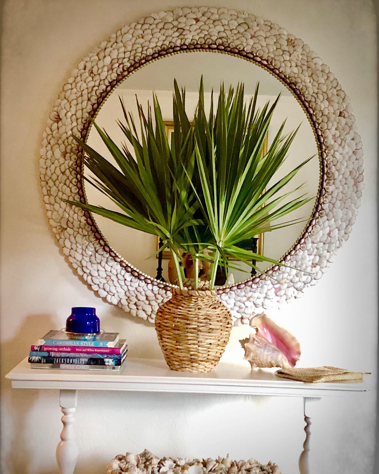 Green palm fronds in a rafia covered vase sitting on a white console table. The table also has books and a shell. A large round shell mirror hangs on the wall behind them.