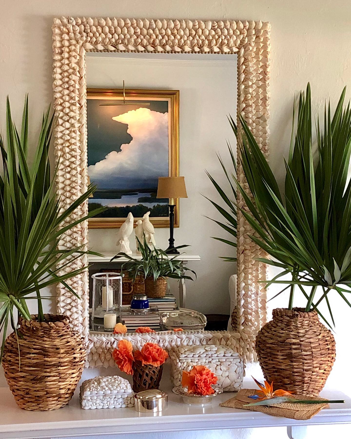 Palm fronds in baskets in front of a large mirror made with white shells. orange hibiscus on the table below it.