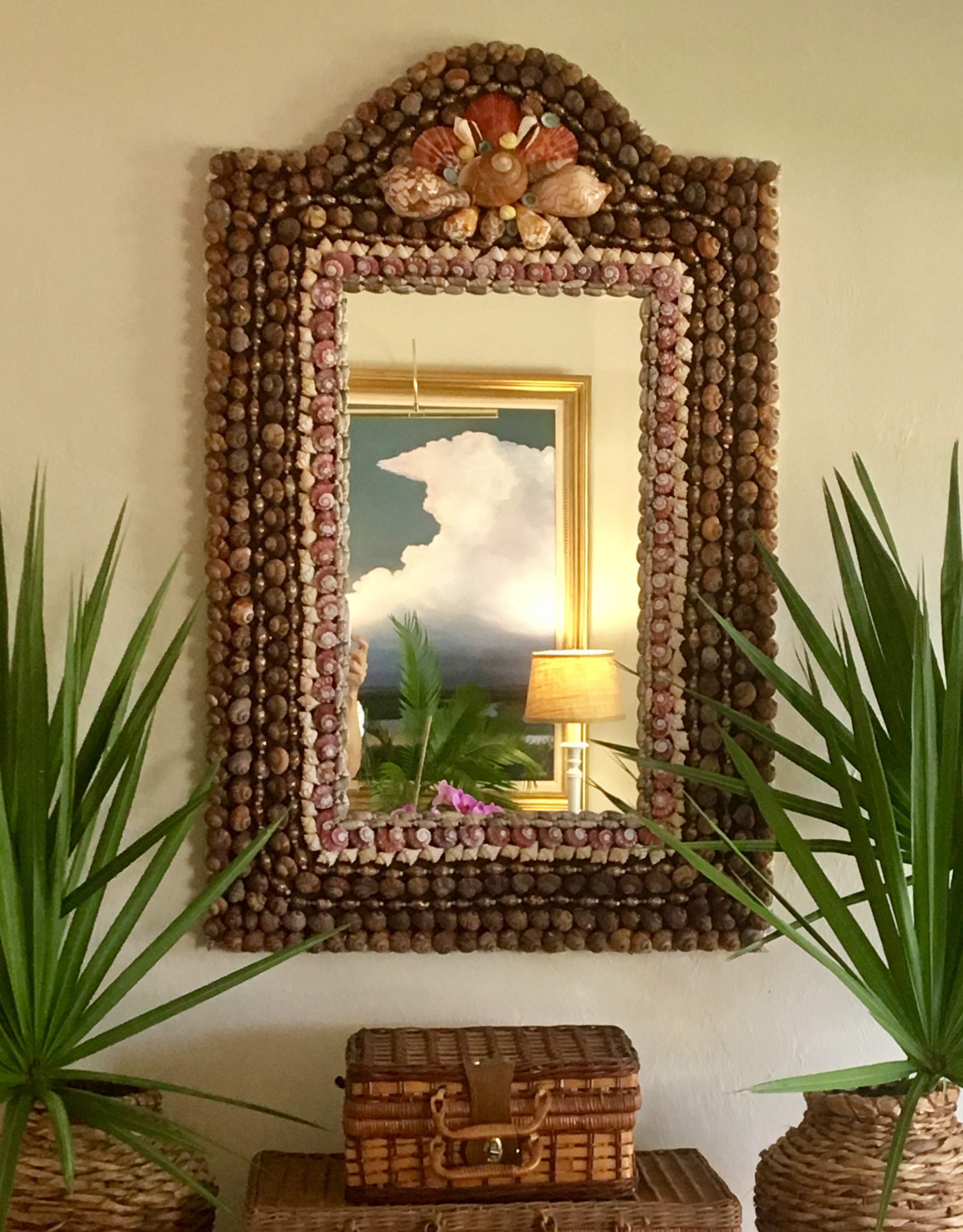A rectangular mirror made with brown and red shells hangs on a wall with a painting with clouds refits in the mirror.