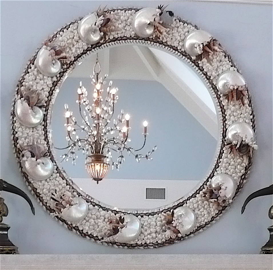 Very large seashell mirror made with white pearl nautilus and other small white shells hangs on a light blue wall with a lit chandelier in it's reflection.