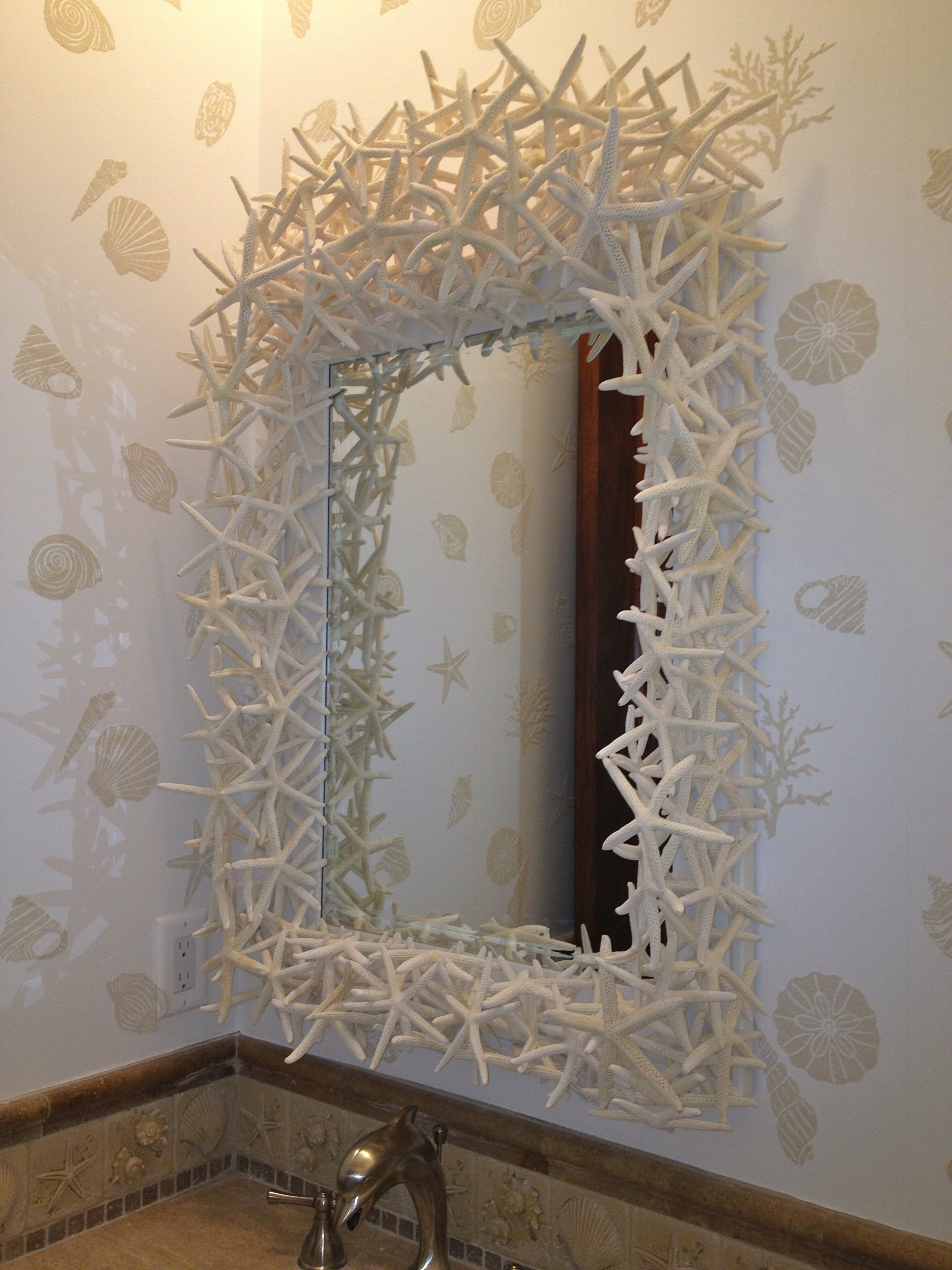 Mirror made with white starfish hangs on a wall with white wall paper with tan shells on it.