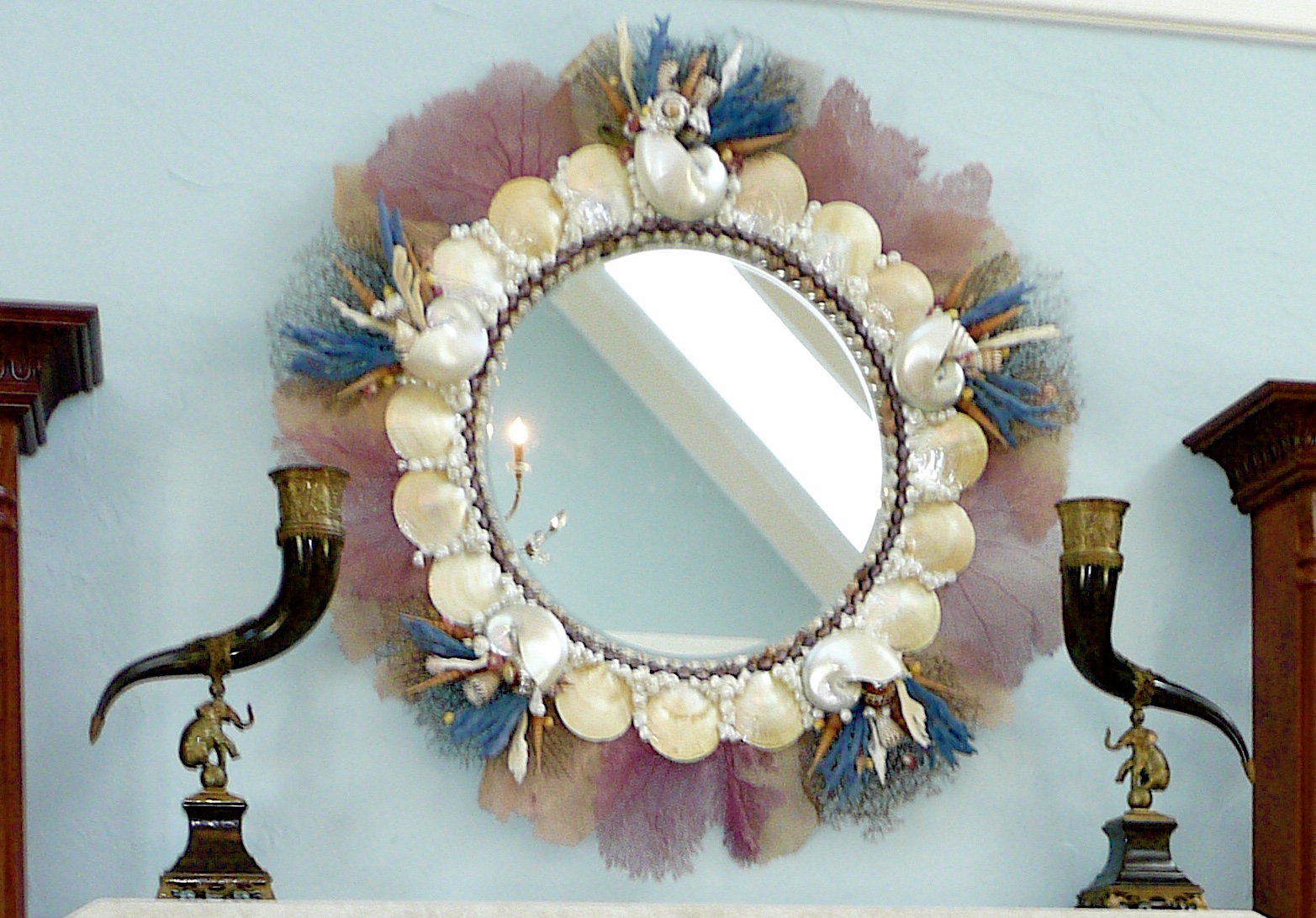 Huge round mirror hanging on a light blue wall. The mirror is made with seashells and large pieces of pink and blue coral protrude all the way around the edges.