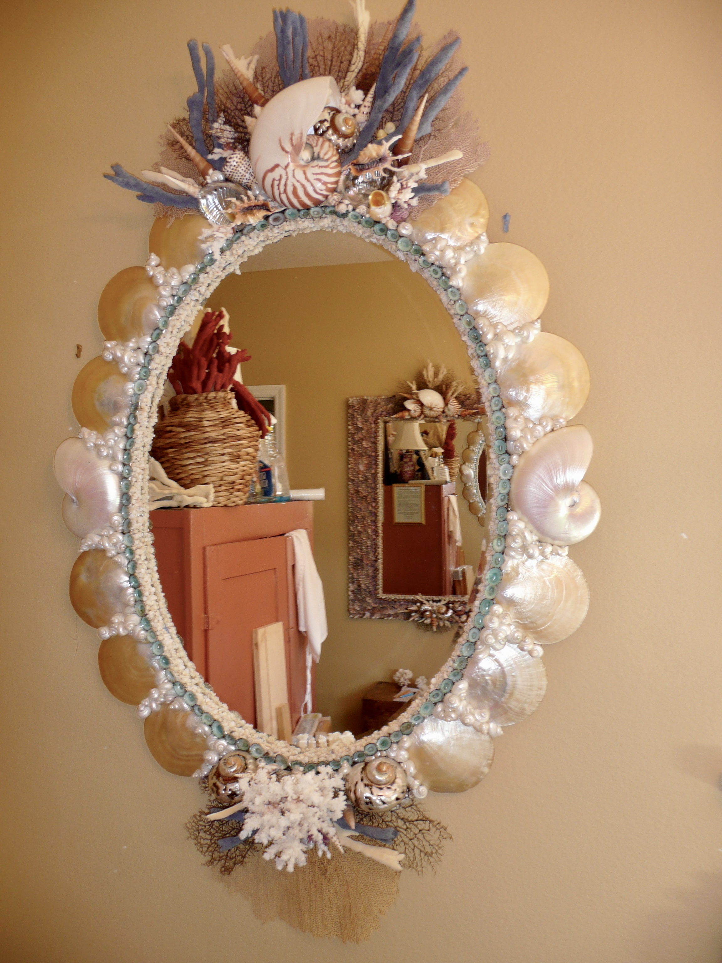 Oval mirror made with large mother of pearl shells is trimmed with row of small green shell on the inside edge. It reflects rust colored furniture and another shell mirror
