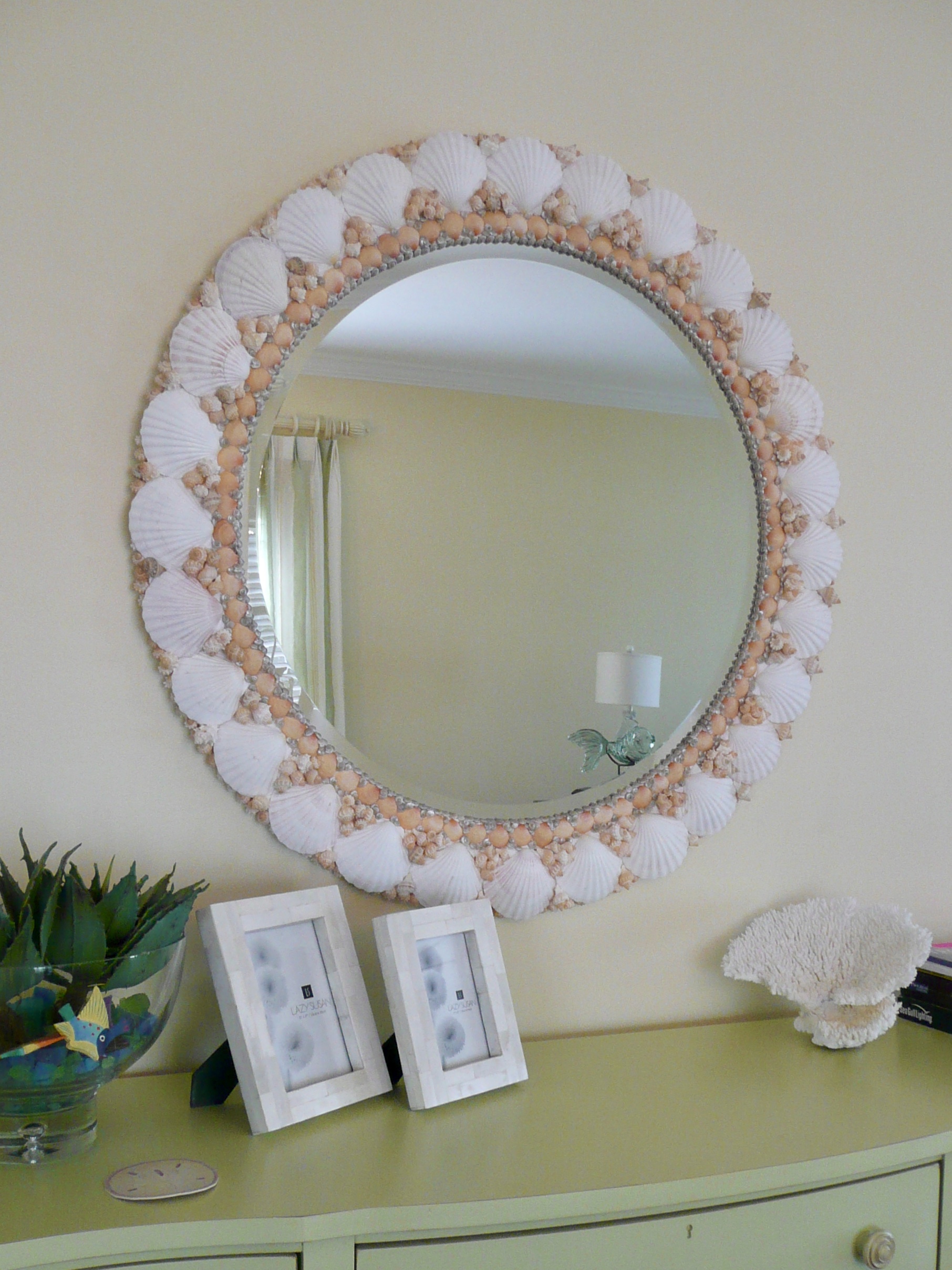 Large round shell mirror with white shells trimmed with peach colored shells along the inside edge hangs over a buffet table painted green.