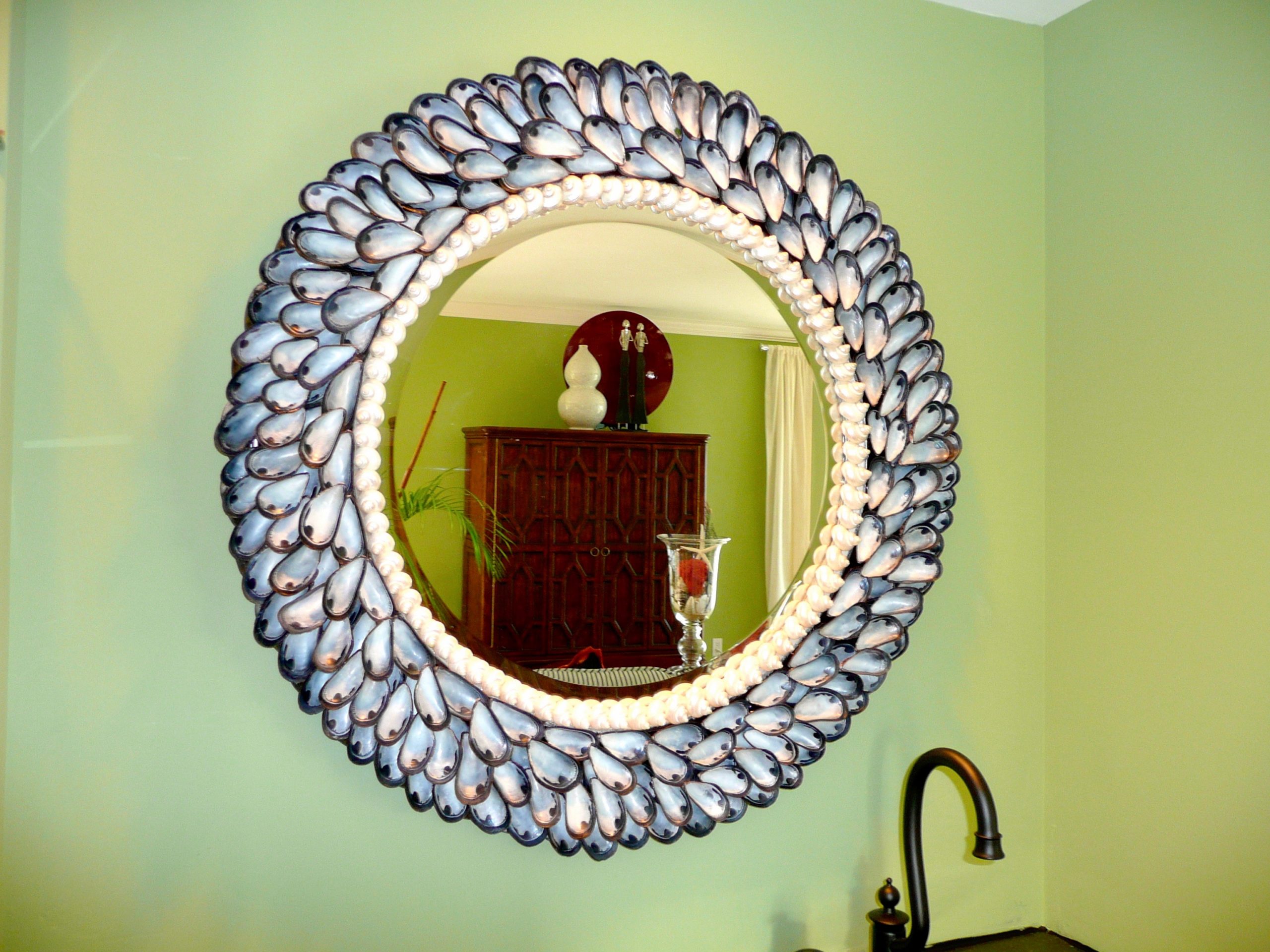 A round shell mirror made with mussel shells on a green wall reflecting an armoire