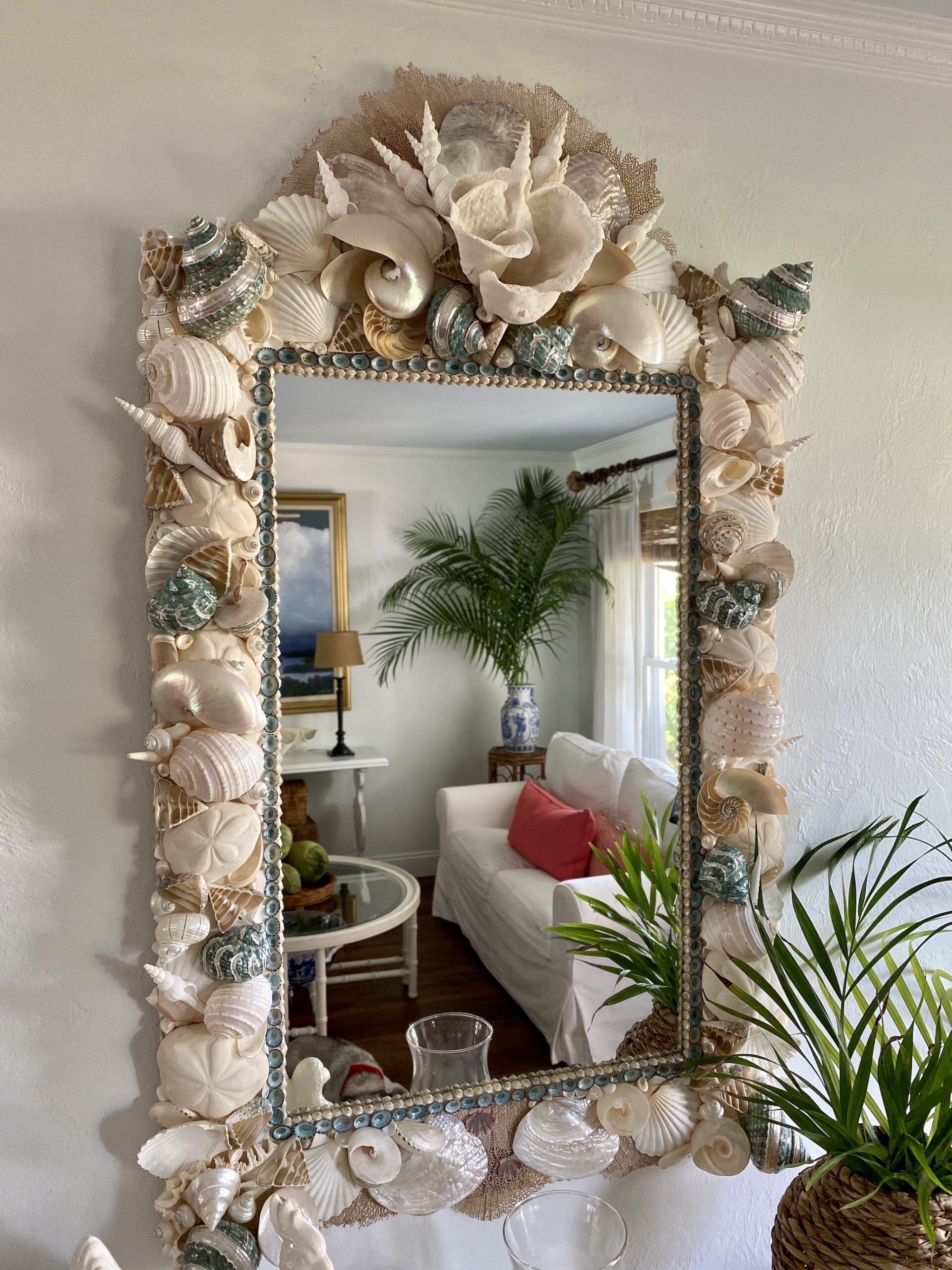 Large shell mirror made with mostly white shells and coral with turquoise shells used as accents. Palm tree reflected in the mirror