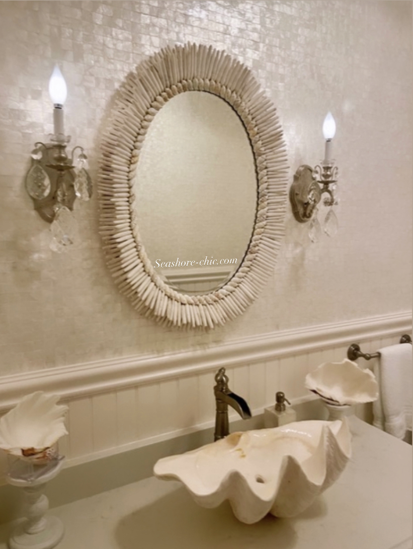 A glamorous all white bathroom with sparkling wall paper has a shell mirror hanging over a sink made from a real clamp shell. Sconces flank each side of the mirror.